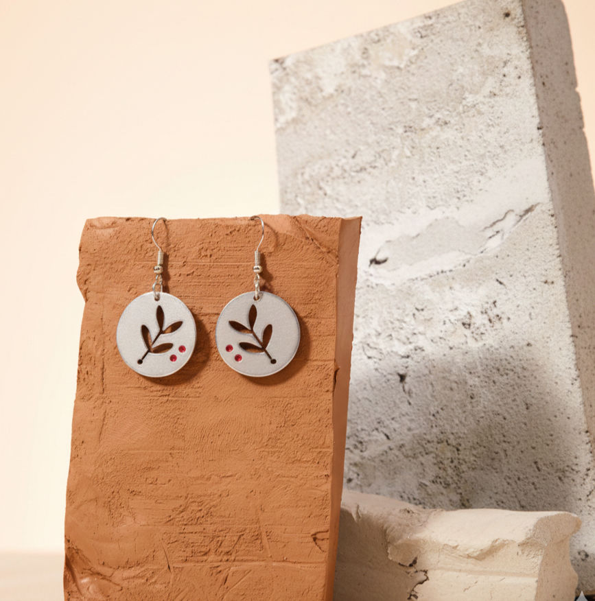 Pendientes de aluminio con forma de rama de olivo colocados junto a hojas reales sobre una superficie de madera, enfatizando