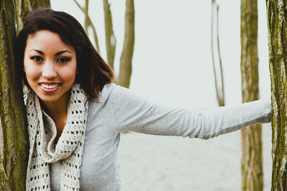 Woman smiling in a gray sweater and knit scarf, leaning between tree trunks on a foggy day. Mood is cheerful and relaxed.