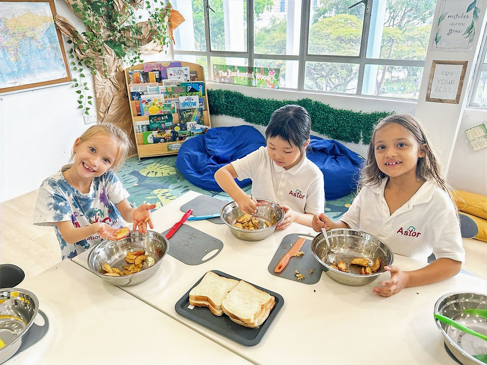 Three young girls are sitting at a table, engaged in a cooking activity at Astor International School. The girl on the left is smiling brightly and holding a piece of food with her hands covered in ingredients. The two girls on the right are mixing food in metal bowls. The setting includes a cozy, well-decorated classroom with bookshelves, a large map on the wall, and encouraging posters, such as "mistakes help you grow" and "try your best." Sandwich bread and cutting boards are visible on the table, indicating they are likely preparing sandwiches or a similar dish.