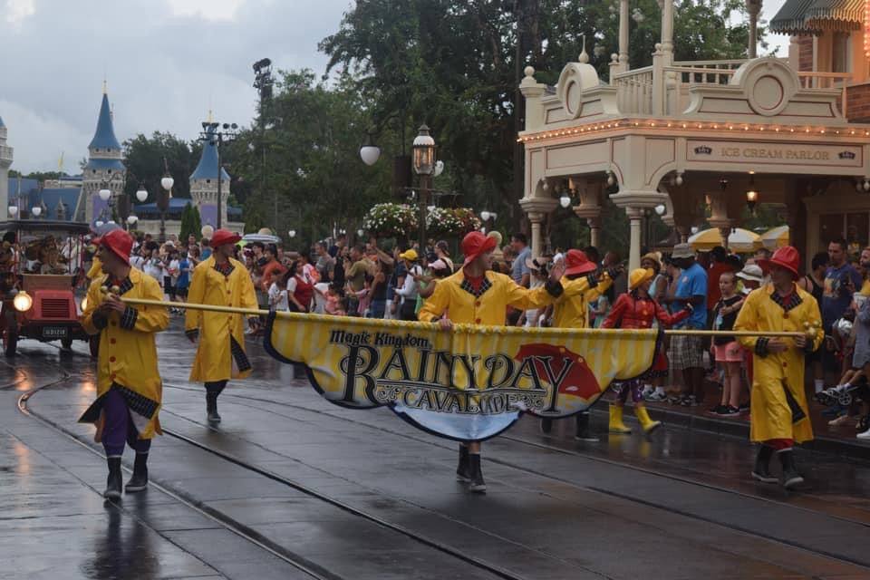 Performers in bright yellow raincoats and red hats lead the Magic Kingdom Rainy Day Cavalcade parade down Main Street, U.S.A. at Walt Disney World during a rainy day, with Cinderella Castle visible in the background and crowds watching under umbrellas.