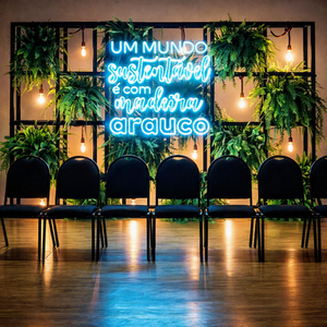 ambiente com cadeiras enfileiradas atrás uma estante modular com lâmpadas e samambaias e placa neon escrito “um mundo sustentável é com madeira arauco”