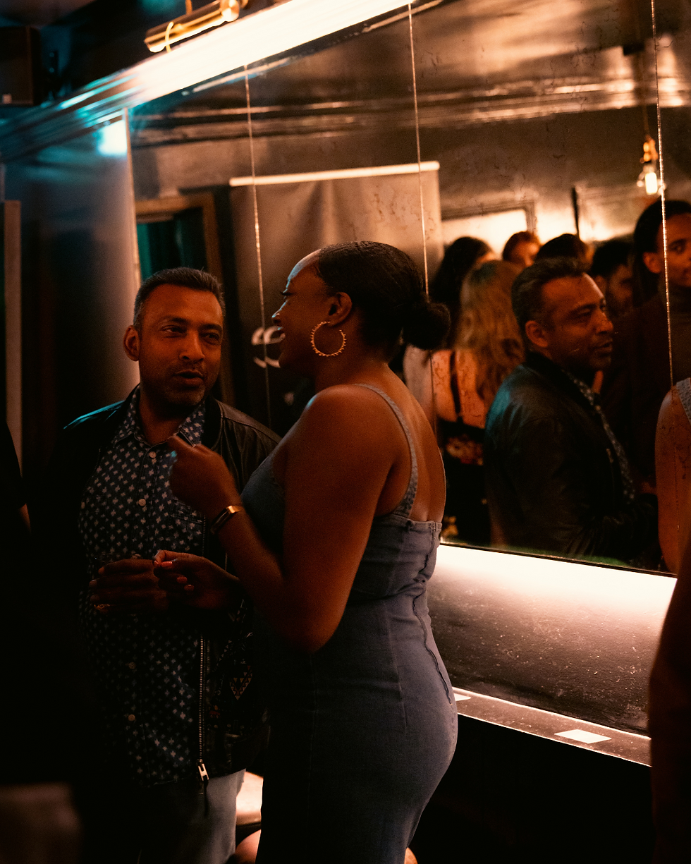 Two people chat animatedly in a dimly lit bar. Mirror reflects more people. Warm lighting creates a lively atmosphere.
