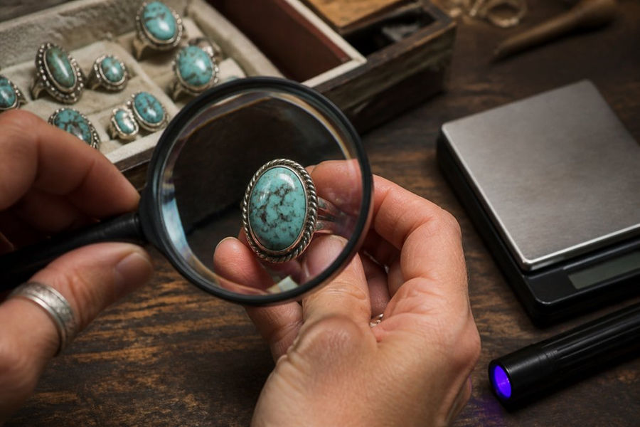 Close-up of hands holding a vintage turquoise ring under a magnifying glass beside a UV pen light and a small digital scale, with a soft jewelry workbench background