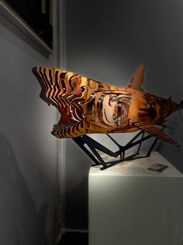 Sculpture de tête de requin métallique or sur piédestal blanc bouche ouverte.