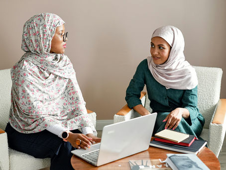 Two women in hijabs sit in an office, engaged in conversation. One types on a laptop, the other holds a notebook. Neutral background. Blog about understanding the realitybehind mental health conditions. 