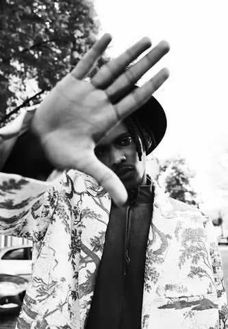 Black and white fashion portrait of a man in a patterned coat and wide-brimmed hat, styled by Stil-ch – Hair Coloring & Fashion Styling Berlin – captured mid-gesture with his hand reaching toward the camera, creating a bold and expressive visual statement.