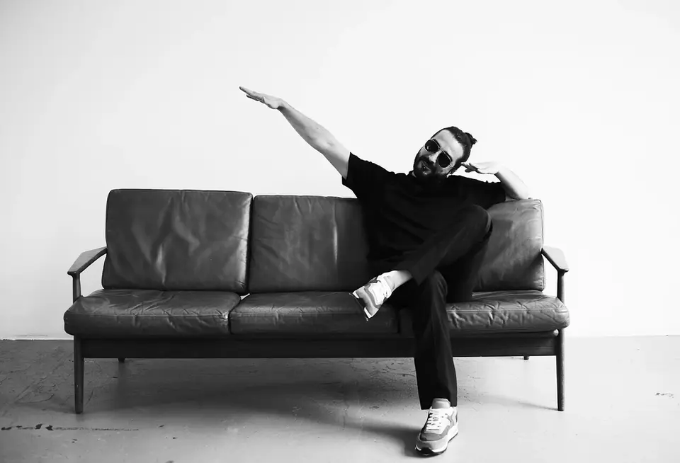 "Stylish man in sunglasses and casual black outfit striking a playful pose on a minimalist leather sofa, with modern grooming and look by Stil-ch – Hair Coloring & Fashion Styling Berlin – captured in a sleek black-and-white editorial setting.
