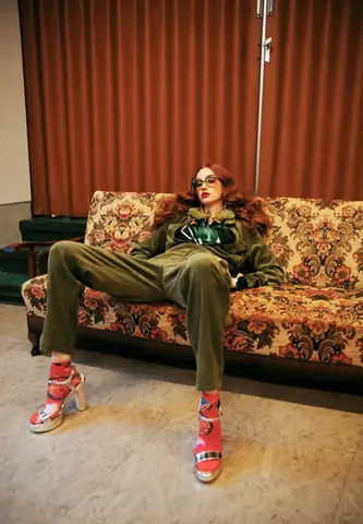 Fashion model styled by Stil-ch – Hair Coloring & Fashion Styling Berlin – lounging confidently on a vintage floral sofa in an olive green corduroy suit, red patterned socks, bold silver platform heels, and retro sunglasses, creating a bold clash of textures and eras.