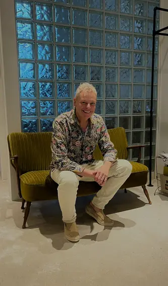 "Smiling man with stylish platinum blonde hair, wearing a colorful floral shirt and light trousers, seated on a retro olive-green sofa in a modern interior – hair and look by Stil-ch, Hair Coloring & Fashion Styling Berlin – with a glass block wall and minimalist decor in the background.