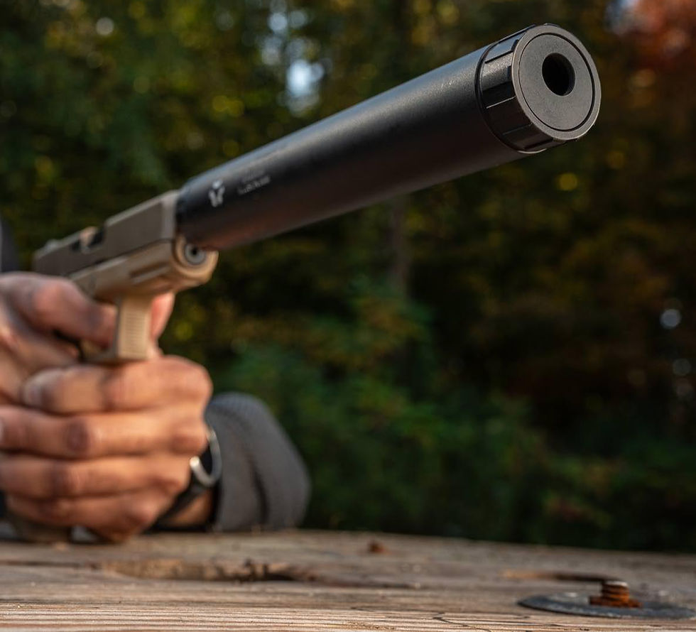 Close-up of a hand holding a suppressed handgun over a wooden table, with blurred green trees in the background.