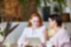 Two women collaboratively viewing a tablet, one gesturing supportively indoors.
