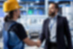 Factory worker and manager shake hands in front of industrial equipment.