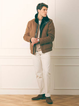 Man in brown jacket and white pants stands indoors with neutral background.