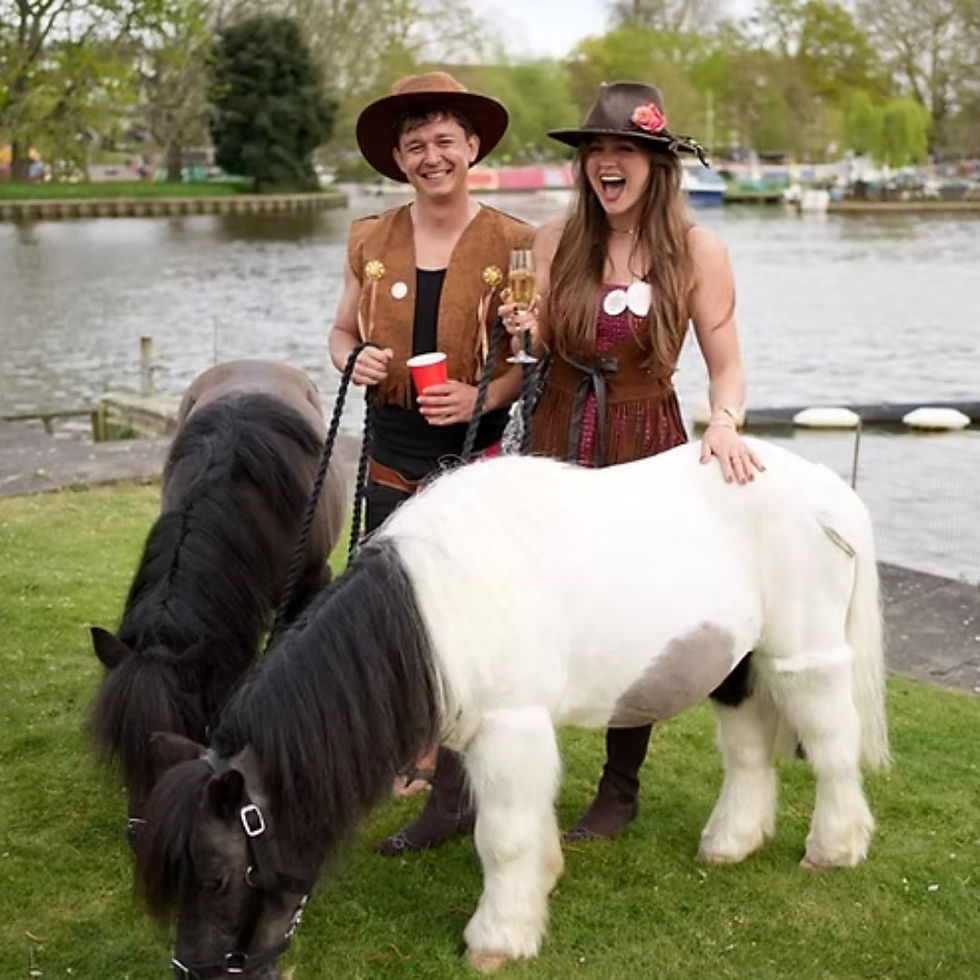 Two people pose with two ponies near a river; celebration, outdoor event.