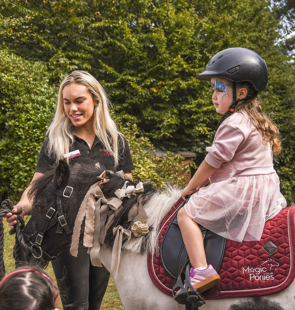 Girl riding pony with Magic Ponies saddle, attendant nearby, outdoors with trees.