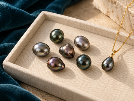 Illustration de perles de Tahiti aux formes et reflets variés, avec un pendentif en or sur un plateau ivoire et un tissu bleu paon.