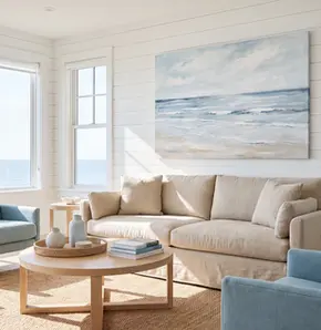 Sunlit coastal living room with oversized blue ocean canvas wall art above a beige sofa, pale blue armchairs, white shiplap walls and ocean-view windows.