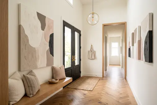 Warm transitional foyer opening into a long hallway decorated with unframed neutral abstract gallery-wrapped canvas art, featuring a large beige, taupe and charcoal statement canvas above a natural wood entry bench and coordinated vertical canvases extending along the corridor beside a matte-black front door.