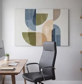 Bright professional home office featuring a large blue, olive, beige and warm-gray geometric gallery-wrapped canvas centered behind a black ergonomic desk chair, with a natural-wood workstation, desktop computer, floating shelves and soft window light creating a polished video-call background.
