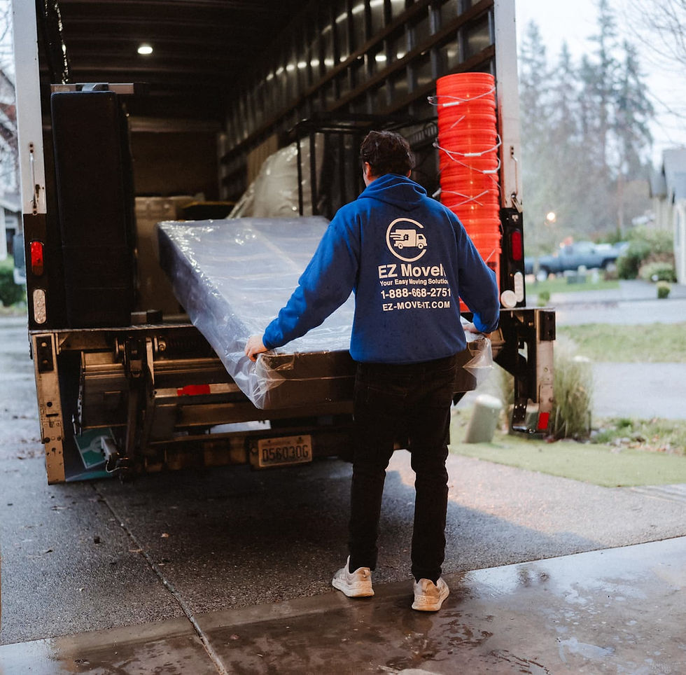 Mover in a blue sweatshirt loads a wrapped item onto a truck. "EZ MoveIt" logo and contact are visible. Overcast day in a suburban area.