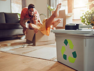 A happy couple having fun packing moving boxes, with a green recycling bin in the foreground, representing eco-friendly decluttering before a relocation.