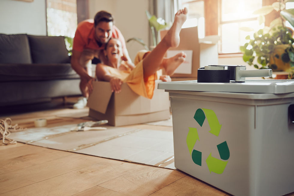 A happy couple having fun packing moving boxes, with a green recycling bin in the foreground, representing eco-friendly decluttering before a relocation.