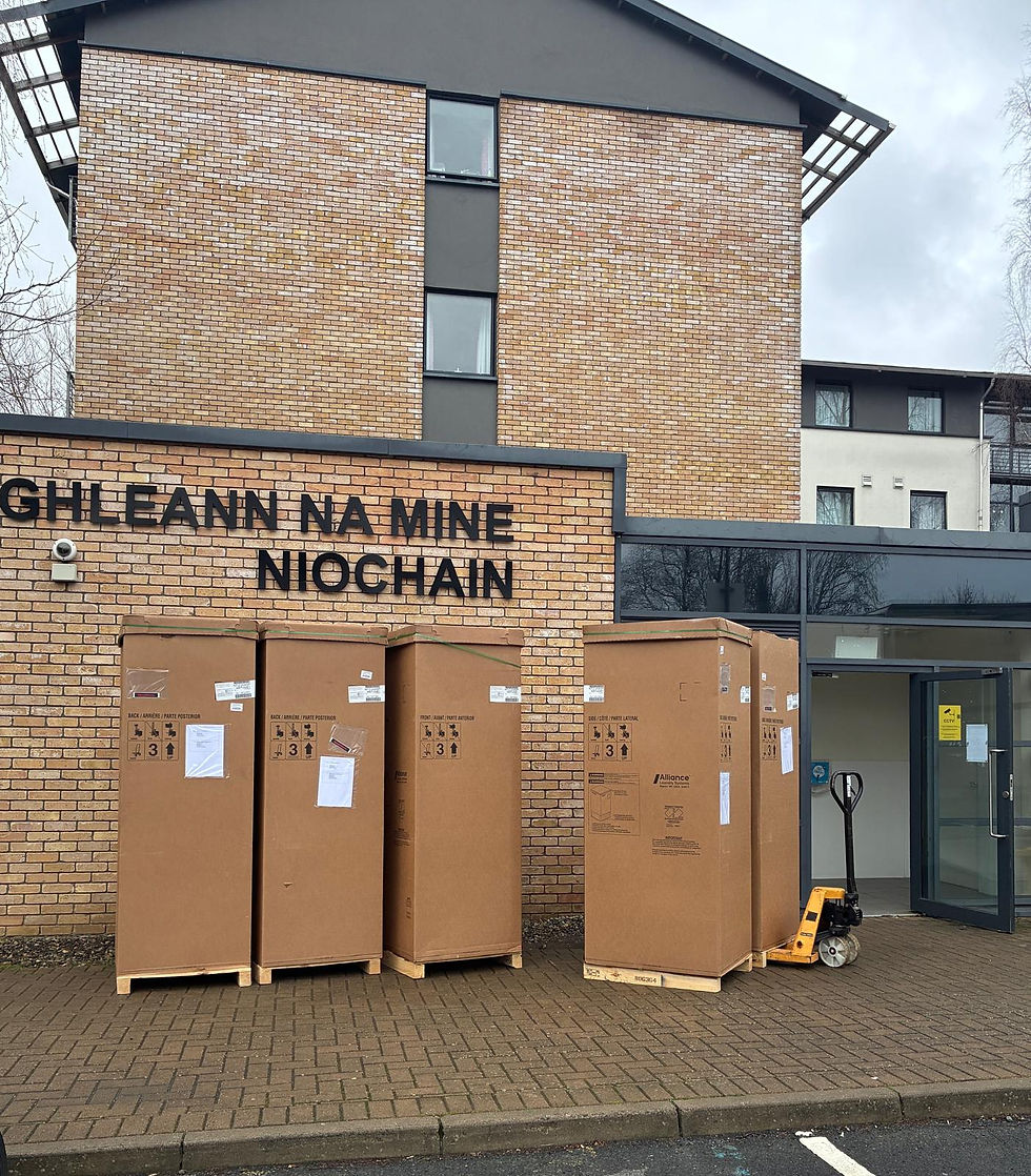Cardboard boxes stacked  laundry machines on a paved path in front of a brick building with signage. A hand truck is nearby, and the sky is overcast.