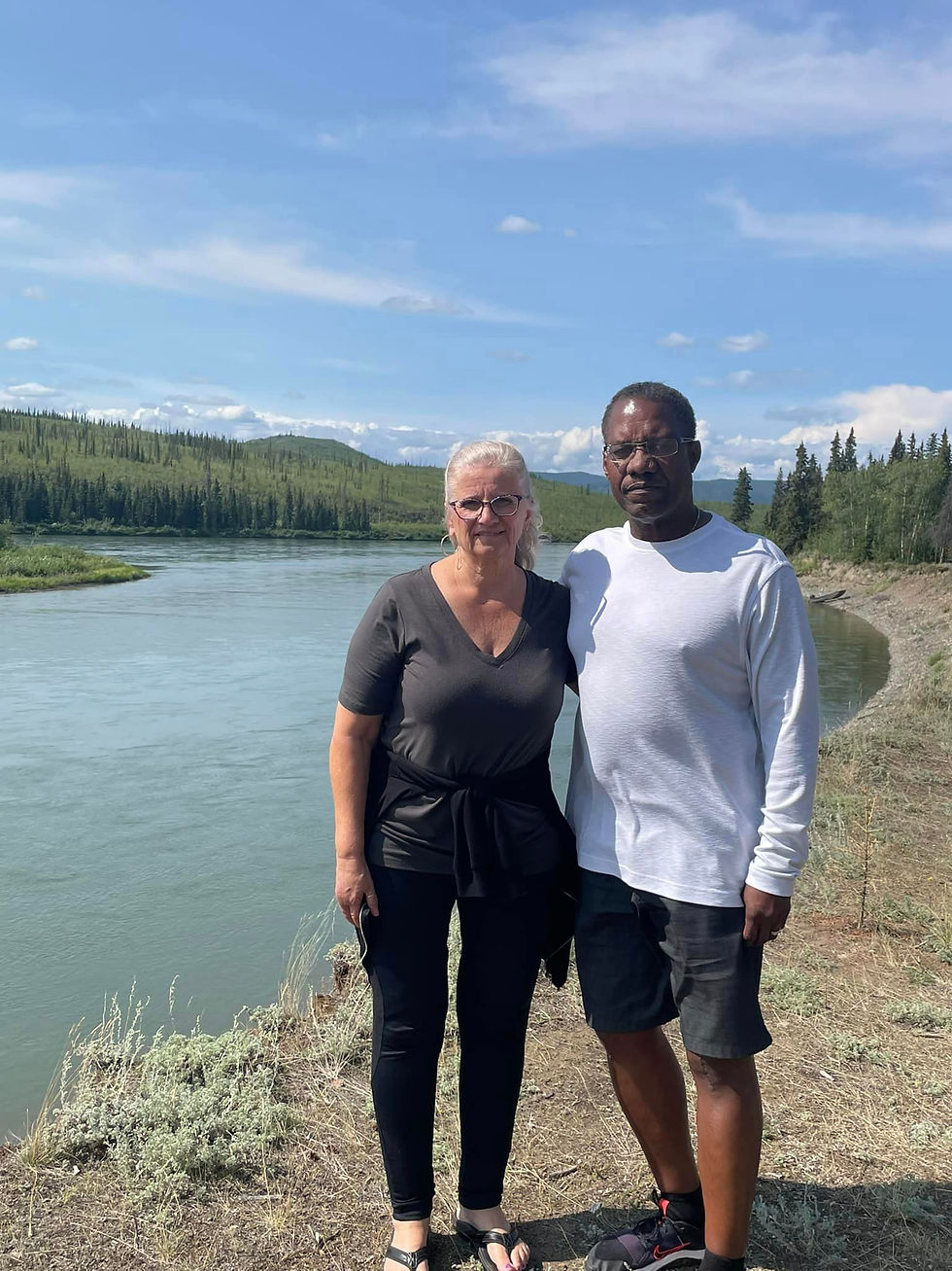 A couple standing together outdoors by a calm river, with a scenic background of forested hills and clear skies. They are dre