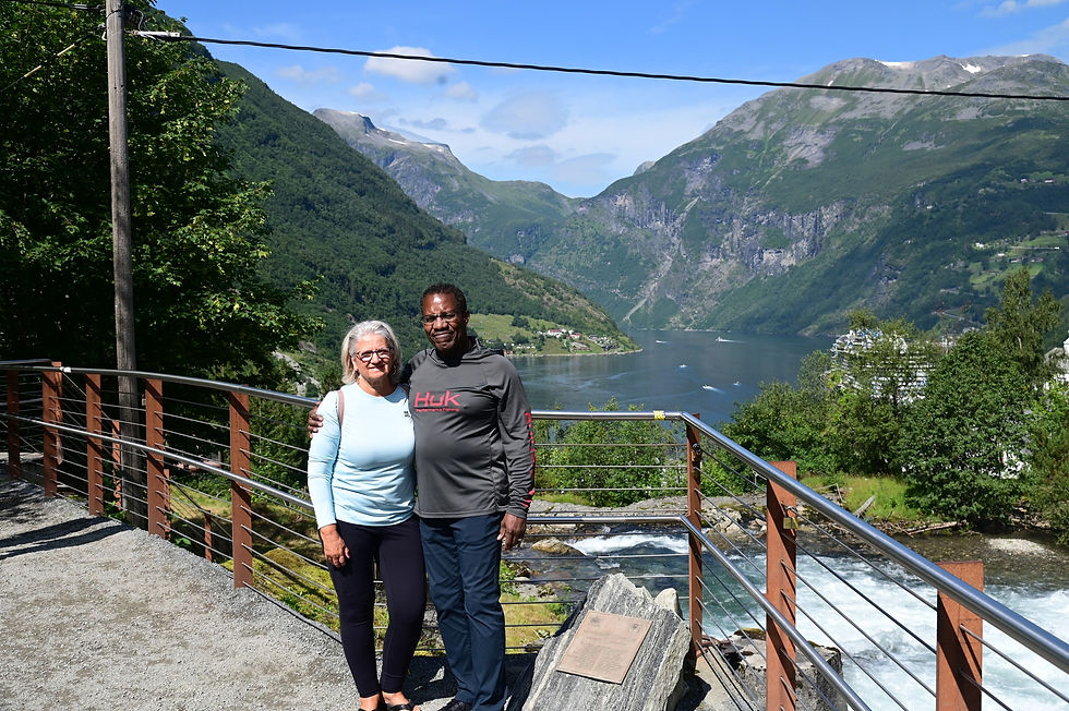 A couple posing together outdoors with a scenic backdrop of mountains, a lake, and lush greenery. They are standing on a path