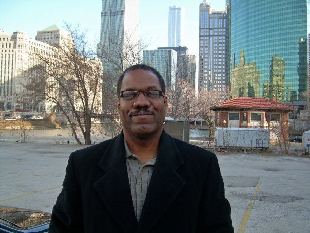 Man wearing a dark overcoat and glasses standing in front of an urban skyline with tall buildings and bare trees, possibly in