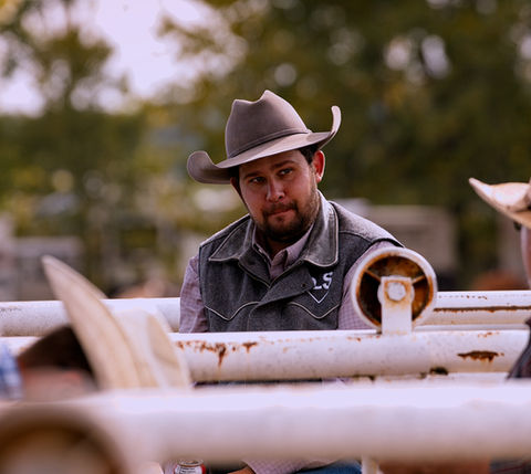 Two cowboys talking, wearing hats, and standing near a metal fence outdoors.