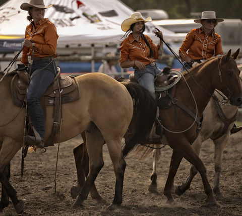 Three cowboys on horseback, wearing orange shirts and hats, riding together. Western Cowboy Chronicles.