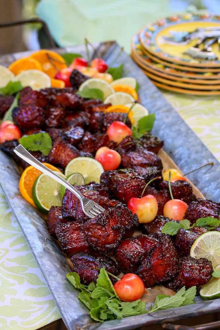 A tray of pork belly burnt ends with limes, tangerines, and cherries.