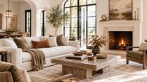 Bright finished living room with a cream sofa, olive upholstered chairs, large wood coffee table, stone fireplace, arched black windows, layered lighting, and warm seasonal accents.
