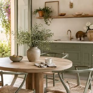 Sunlit rustic kitchen with open door, round wooden table, sage chairs, potted greenery, and brass sink in a calm, airy room.
