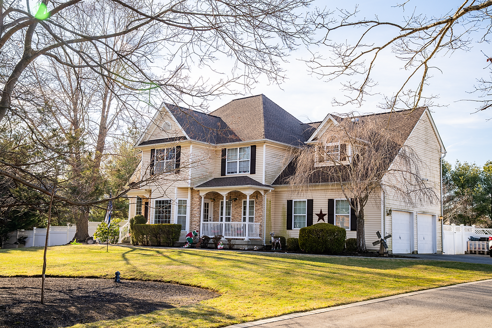 Two-story suburban house with beige siding, black shutters, and a gable roof. Surrounded by trees and a well-manicured lawn under a clear sky.