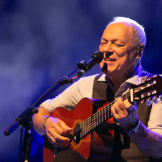 Toquinho se apresenta ao vivo cantando e tocando violão no palco, durante show iluminado em tons de azul.