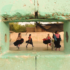 Cena de “Bailarinas de San José”, com grupo de mulheres dançando ao fundo, vistas através de uma abertura retangular em uma parede verde desgastada.