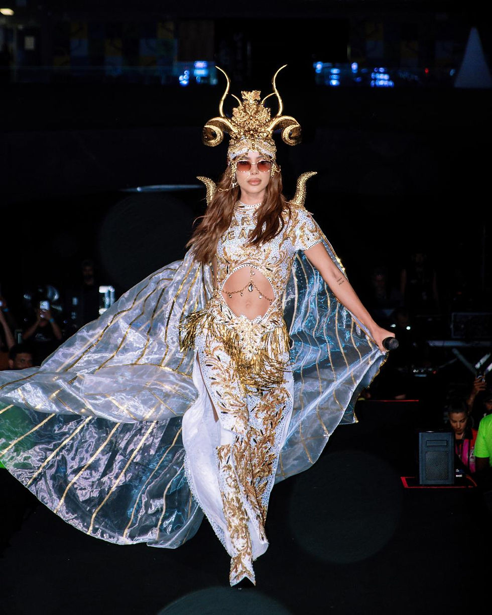 Anitta se apresenta nos Ensaios da Anitta usando figurino branco e dourado com adereço de cabeça ornamentado e capa translúcida, enquanto segura um microfone no palco.