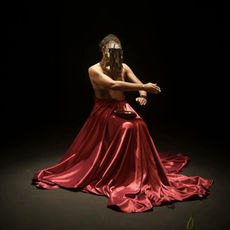 Cena do filme “Segunda Pele”, com performer usando vestido vermelho e rosto coberto por adereço dourado, sentada sob foco de luz.