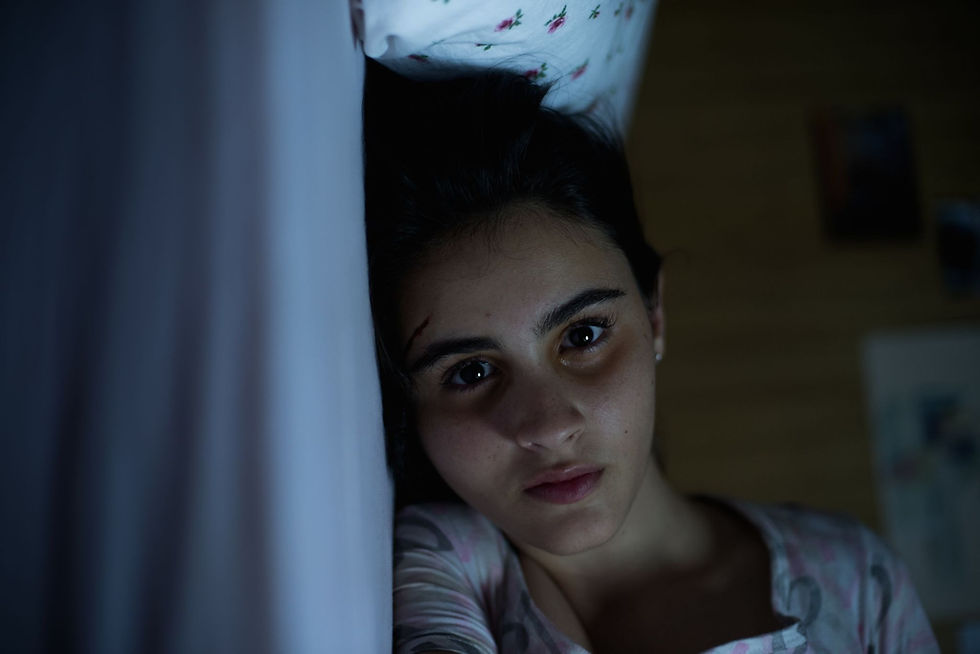 Jovem deitada em uma cama observa diretamente para a câmera em ambiente com pouca iluminação, em cena do filme Mate-me Por Favor.