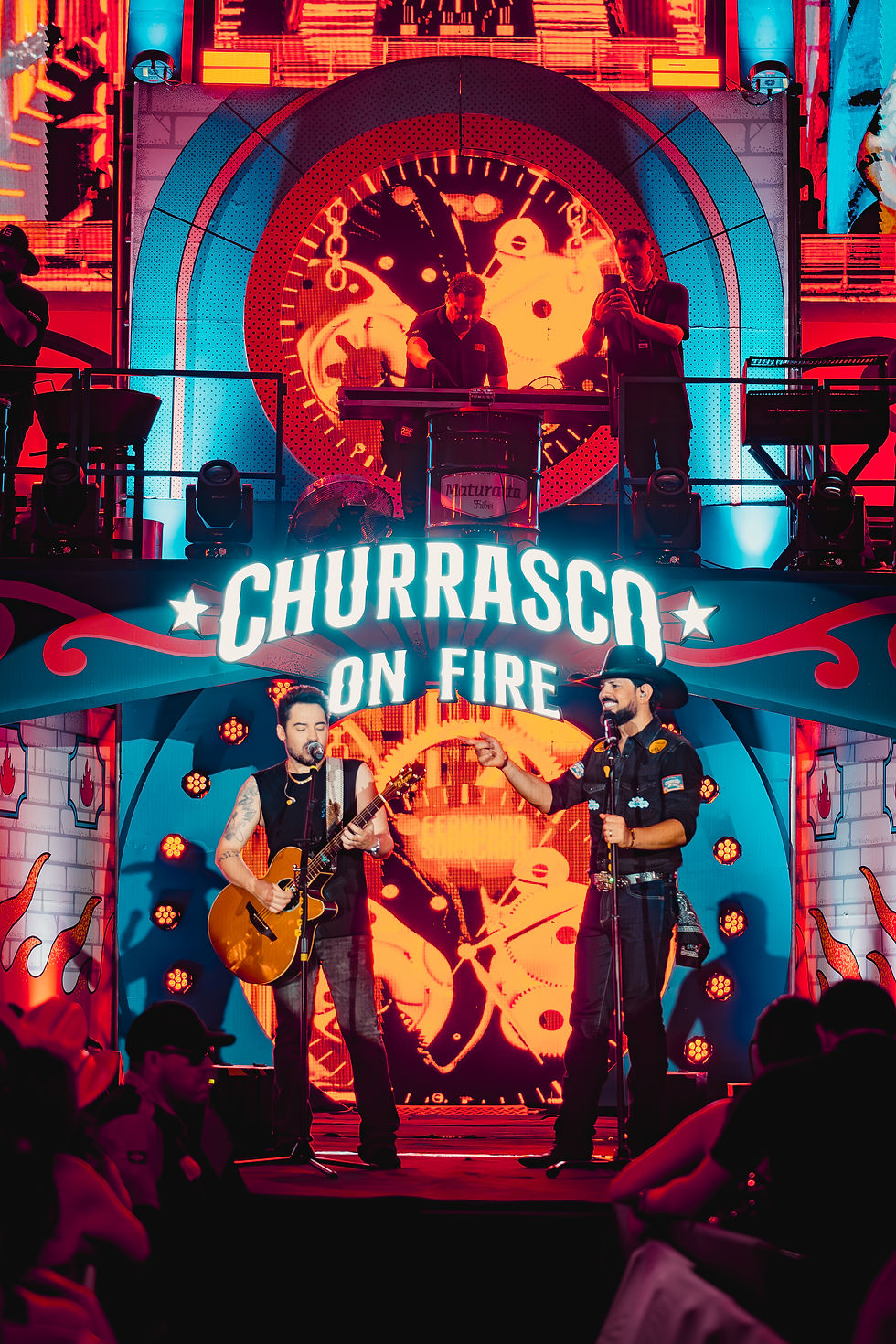 Dupla Fernando e Sorocaba se apresenta no palco do Churrasco On Fire, com iluminação em tons vibrantes, telões de LED ao fundo e o público acompanhando o show. A estrutura do evento destaca a marca “Churrasco On Fire” no cenário principal.