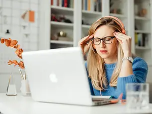 Woman in glasses with headphones looking stressed, holding her head in hands, sits at a desk with a laptop.
