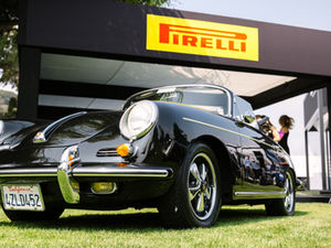 im Vordergrund ist ein schwarzer Porsche mit 911er Pirelli-Reifen (etwas von unten fotografiert, auf einer Wiese stehend), im Hintergrund - direkt über dem Auto - ist das gelb-rote Logo von Pirelli zu sehen