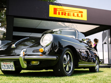 im Vordergrund ist ein schwarzer Porsche mit 911er Pirelli-Reifen (etwas von unten fotografiert, auf einer Wiese stehend), im Hintergrund - direkt über dem Auto - ist das gelb-rote Logo von Pirelli zu sehen
