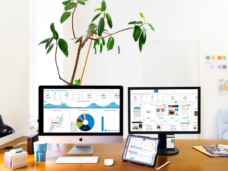 auf einem Holz-Schreibtisch in einem Büro stehen mittig 2 große Bildschirme (mit einer Tastatur und einer PC-Maus), 1 Tablet und 1 Handy, die unterschiedliche Programme mit Statistiken zeigen; a, Schreibtisch sind links noch eine schwarze Schreibtischlampe, ein Lautsprecher, ein Kaffeebecher, ein Notizblock und ein Marker zu sehen, rechts liegen ein paar Prospekte; im Hintergrund ist eine Pflanze zu sehen und ein paar Skizzen auf Blättern, die auf der Wand hängen