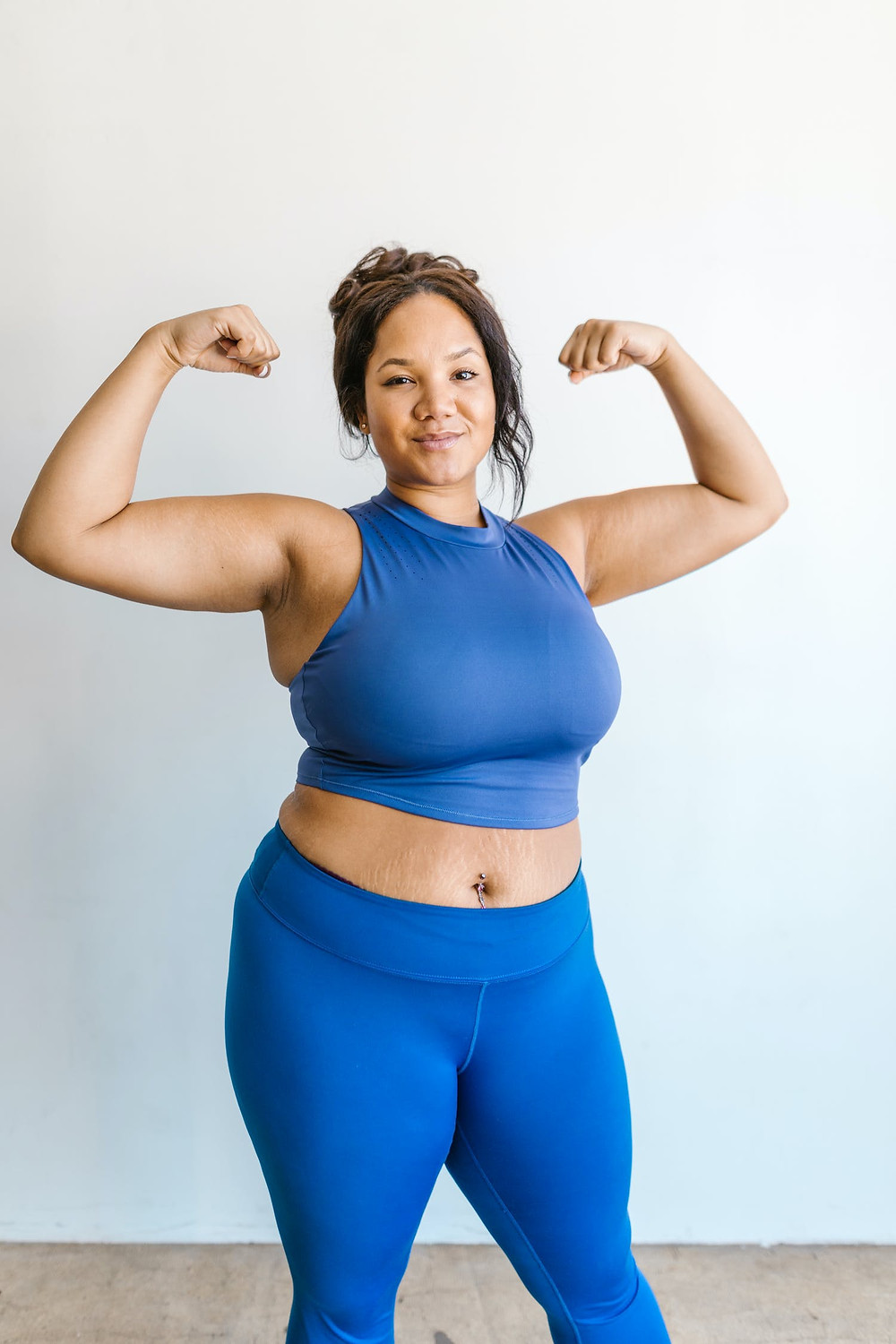 Fat brown mixed race woman in blue athletic crop top and leggings proudly flexing her biceps and smiling