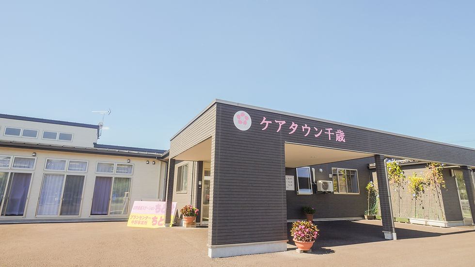 青空の下、黒と白の建物の玄関にピンクの看板「ケアタウン千歳」。鉢植えの花が並ぶ静かな施設外観】【。