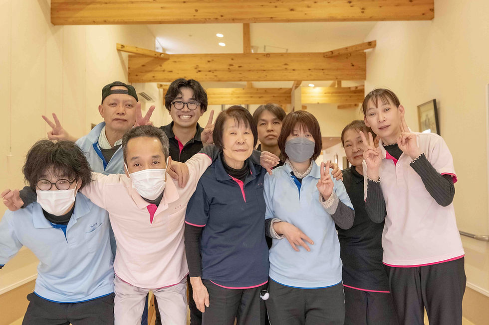 室内で8人の人々がポーズをとり笑顔を見せる。木製の天井が特徴的で、ピンクや青いシャツを着用。和やかな雰囲気。