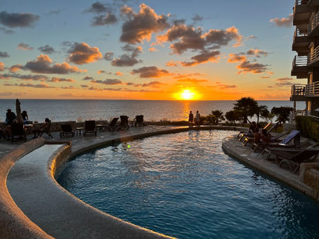 Cupecoy Bay resort-style pool at sunset, with lounge chairs, a few people by the water, and a glowing orange sky over the calm sea.
